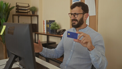 Hispanic man with beard indoors in office examines credit card, appearing thoughtful and concerned at computer desk, showcasing a professional and analytical environment.