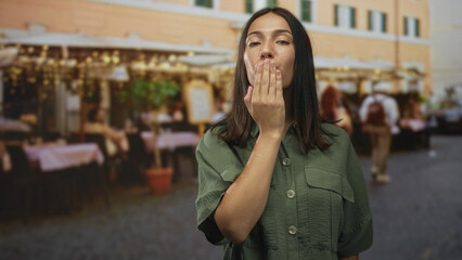 Woman blowing a kiss with hands to lips at a street cafe terrace near tables and lights; romance playful warmth.