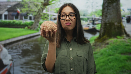 Young brunette woman with glasses holds anatomical brain prop in outstretched hand beside park canal and mossy tree with boat in view; disgust curiosity.