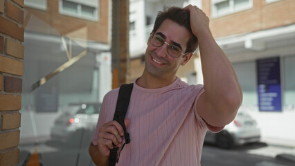 Man smiles, runs hand through hair and holds backpack strap on a sunlit urban street with parked cars; casual ease relaxed.