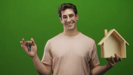 Man holding keys and wooden house model in green studio, smiling and presenting keys with outstretched hand; pride new home.