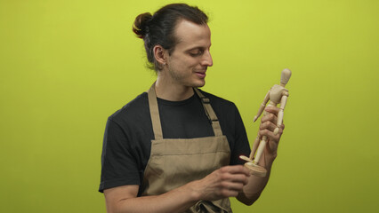 Man holding a wooden artist mannequin and examining it in lime green studio, young hispanic with long hair and apron; creative thinking.