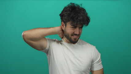 Young hispanic man with beard in white shirt expressing neck discomfort against a green background,...