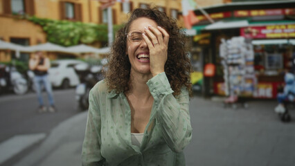 Woman laughing with hand covering face on busy street wearing glasses and a green blouse, curly...