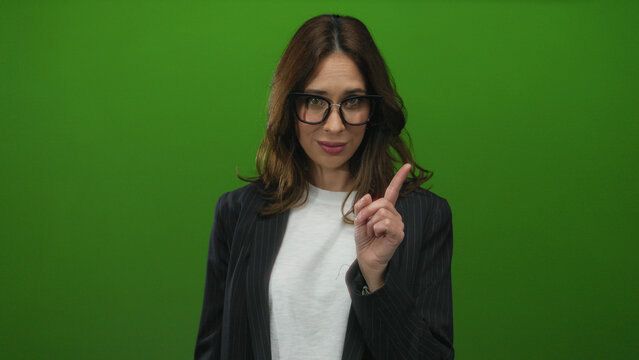 Woman gestures disagreement with finger on green background, showcasing young hispanic female in glasses and black jacket emphasizing refusal and self-aware expression.