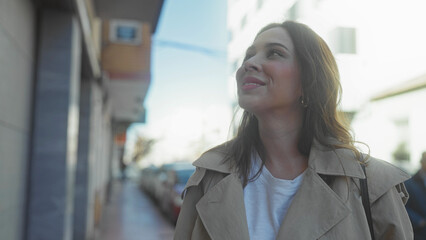 Woman walking on urban street looks up at buildings on a sunny day wearing a trench coat in city.