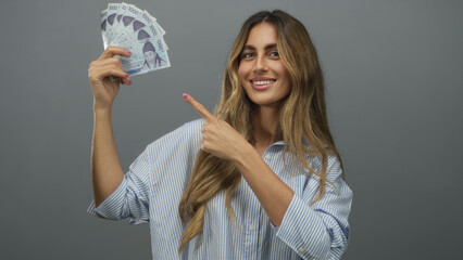 Young blonde woman wearing striped shirt holds korean won banknotes and points finger in studio;...