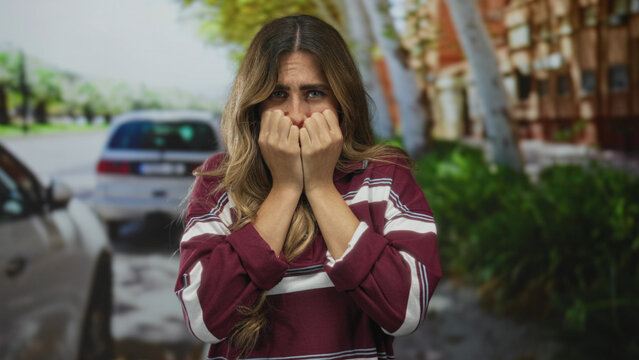Young blonde woman covers mouth with hands and widens eyes on sunlit suburban street; shock fear anxiety.