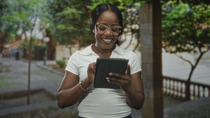 Fototapeta premium Woman holding tablet with fist raised and smiling, wearing glasses and bracelet, standing on street; confidence success.