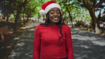 Woman wearing a santa hat and red sweater smiles and looks sideways on a tree lined street with park benches; festive joy.