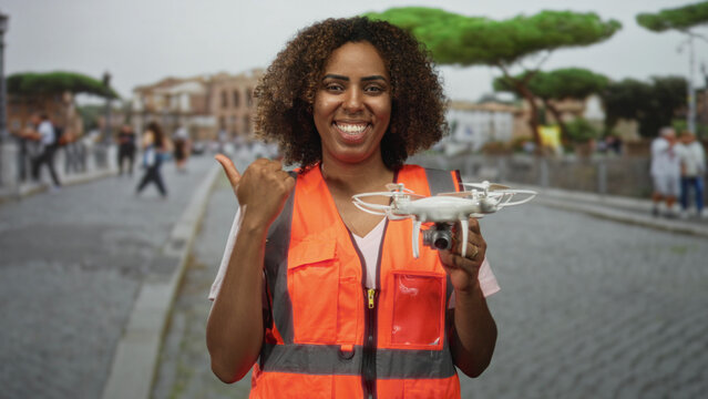 Woman holding drone and giving thumbs up on street wearing orange safety vest; confidence technology empowerment. - Powered by Adobe