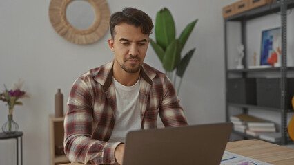 Young man working in an office setting, seated at a desk with a laptop, surrounded by indoor plants and decor, focusing on tasks in an organized, stylish workspace.