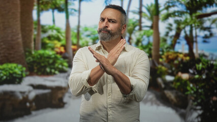 Man middle aged hispanic with grey beard and arms crossed x in park near palms and green foliage; boundary rejection.