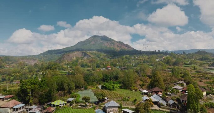 Indonesia, Bali: Mountain Abang volcano rises above a rural Balinese village, lush terraced fields and dense trees, sunlit farmland and traditional homes under a clear blue sky. Aerial rise up flight