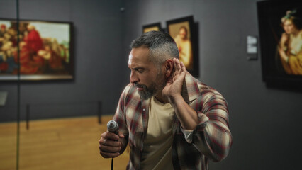 Man holding drill cups hand to ear in gallery beside framed paintings, listening to the tool's sound while examining exhibit for damage; quiet curiosity.