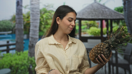 Woman holding pineapple with hands and fingers by hotel pool near building gazebo at resort, inspecting fruit with slight smile; tropical curiosity.
