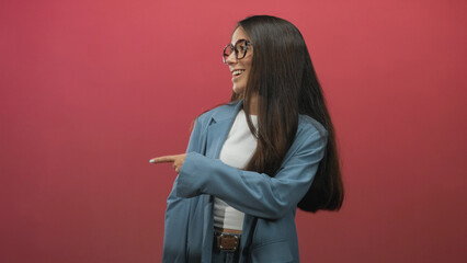Young woman in blue blazer and glasses pointing finger to the right while smiling in studio; joyful confidence.