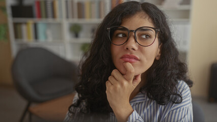 Young brunette woman wearing eyeglasses, hand on chin and finger to lips in an office building; thoughtful curiosity.