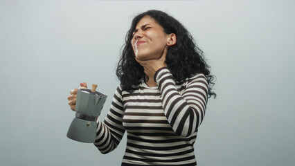 Arab woman holding a moka pot and clutching her throat with one hand, eyes closed and grimacing, wearing a striped shirt in a pale blue studio; distress.