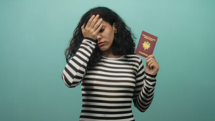 Woman holding passport and covering face with hand to forehead in studio with teal backdrop; travel...