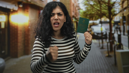 Young arab woman holding saudi passport with clenched fist gesture on a city street with storefronts and trees; frustration resilience.