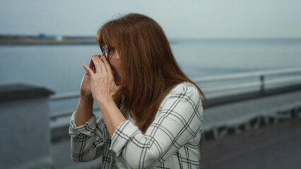 Senior hispanic woman wearing glasses and checkered shirt animatedly shouting near a seaside promenade, capturing outdoor excitement with ocean in the background.