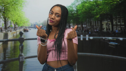 Fototapeta premium Black woman on a canal bridge making finger heart gestures with hands and visible midriff in a pink crop top; playful affection.