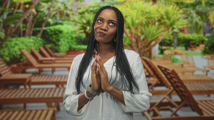 Woman with hands pressed together beside wooden lounge chair in building, wearing white blouse and bracelets; serenity contemplation.