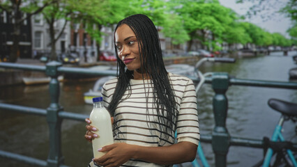 Fototapeta premium Woman holding milk bottle in hand on street by canal, striped shirt and braided hair visible; serenity sustainability.