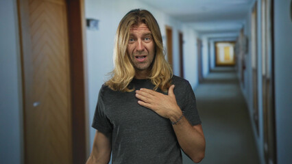 Blond man places hand on chest in a spacious luxury hotel building corridor with wooden doors...