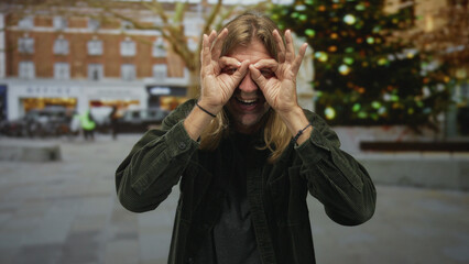 Fototapeta premium Blond man with long hair raises bare hands to form hand binoculars on a busy cobblestone street at midday; watching exploration joy.