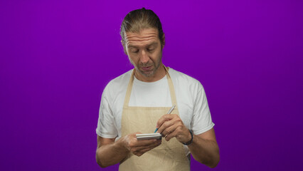 Man with long hair writing in notepad as he holds a pen and looks thoughtful in a purple studio;...