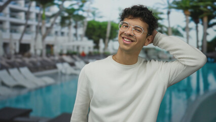 Fototapeta premium Man smiles while tilting head at resort pool adjacent to hotel building with palm trees in view; relaxation.