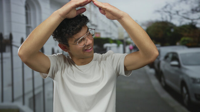 Young hispanic man in white shirt and glasses smiles and forms roof with hands on street; home happiness.
