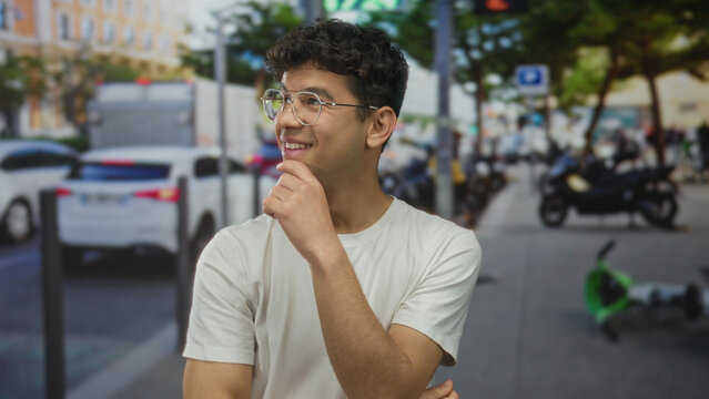 Young hispanic man with hand on chin on a busy city street lined with vehicles and trees looking sideways; quiet contemplation. - Powered by Adobe