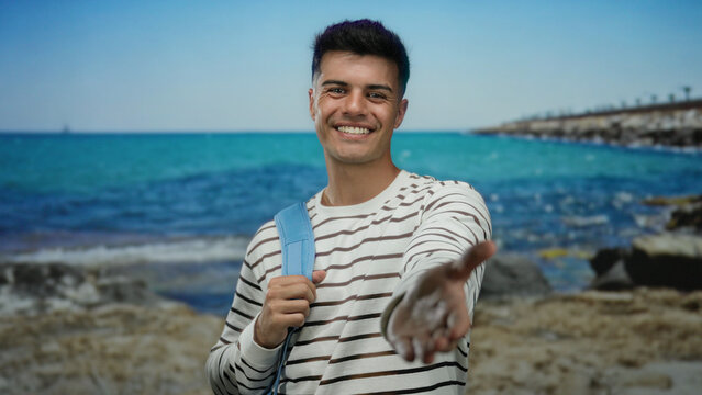 Young man with a striped shirt smiles warmly at the seaside, extending his hand towards the camera, with a turquoise sea in the background, capturing a friendly summer moment.