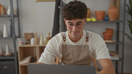 Man adjusts glasses in studio at laptop wearing apron, surrounded by pottery shelves; concentration craftsmanship creativity.