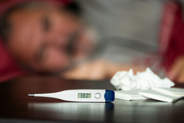 Fever concept with digital thermometer and tissues on a table, sick man resting on a sofa