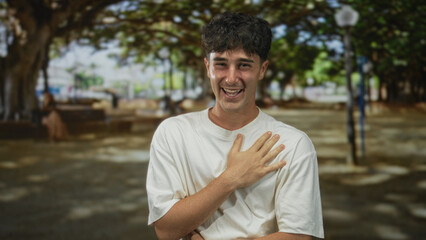 Fototapeta premium Man with hand on chest laughing in a tree lined street, wearing white t shirt and relaxed posture, visible bare hand; joy sincerity warmth.