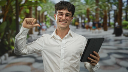 Fototapeta premium Young hispanic man smiling holding tablet with thumb pointing to chest on street lined with palm trees and blurred pedestrians; confidence success.