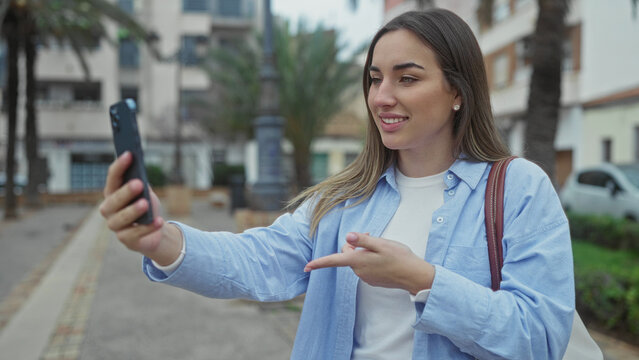 Young woman outdoors on street, using smartphone, gesturing during video call, dressed casually, showcasing urban lifestyle, engaging in digital communication with vibrant backdrop.
