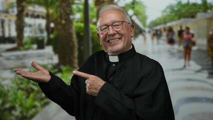 Fototapeta premium Senior priest wearing glasses and black clerical robe happily points at his palm while standing in a lively outdoor street setting with people walking by.