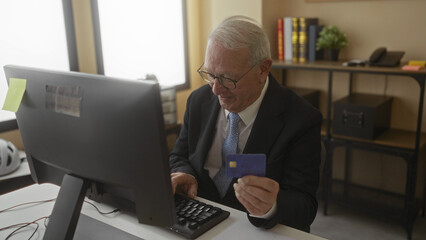 Senior man in business suit using computer in office holding credit card indicating online transaction with smile in professional workplace interior setting