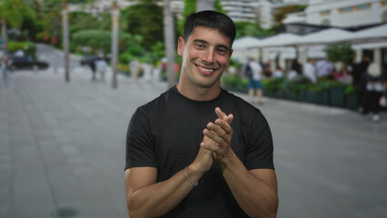 Young hispanic man wearing black shirt clapping hands and smiling on street outdoors; celebration joy.