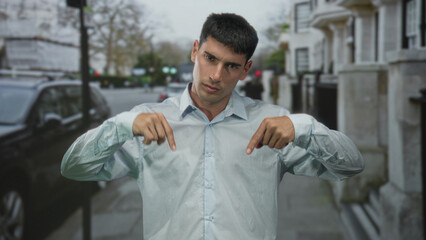 Man in light blue shirt pointing both fingers downward at sidewalk on urban street lined with parked cars and stone buildings; confidence.