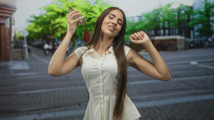 Fototapeta premium Teenage girl in white dress holds smartphone to ear and raises fist while dancing on city street; carefree joy.
