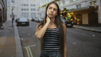 Fototapeta premium Teen girl touching neck and pouting on a city street with parked cars and lit storefronts, long hair and striped tank top; discomfort isolation.