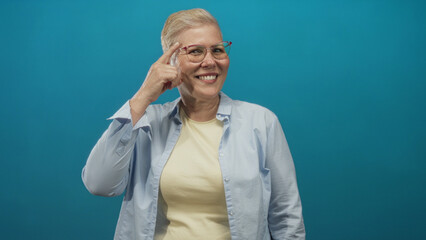 Woman points finger to temple wearing glasses in a studio with a blue backdrop and teal wall emphasizing the thoughtful gesture; confidence wisdom.