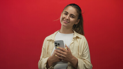 Woman in beige shirt tapping smartphone with yellow nails in studio with red backdrop; concentration.