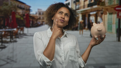 Woman holding piggy bank with hand on chin and pointing finger in thought on street cafe; savings...